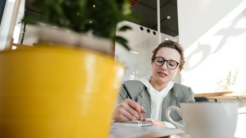 Eine junge Frau in einem Café, die Notizen macht, eingerahmt von einer Topfpflanze und einer Kaffeetasse.