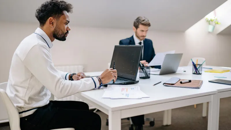 Zwei Männer arbeiten kollaborativ in einem modernen Büroumfeld und konzentrieren sich auf Aufgaben und Laptop-Bildschirme.