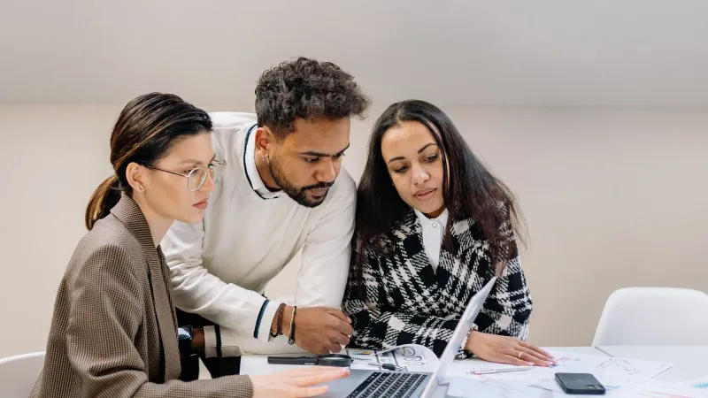 Drei Fachleute, die an einem Tisch mit einem Laptop und Dokumenten zusammenarbeiten, zeigen Teamarbeit und Vielfalt.
