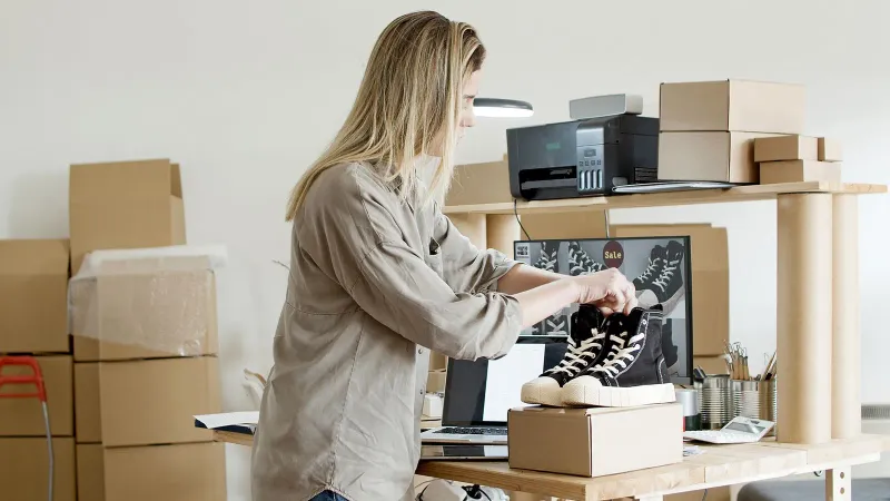 Eine Frau verpackt Sneakers in eine Box an ihrem E-Commerce-Arbeitsplatz, umgeben von Verpackungsmaterialien.