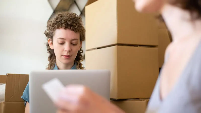 Frauen, die ein kleines Unternehmen führen, umgeben von Kartons und einem Laptop für Online-Bestellungen.