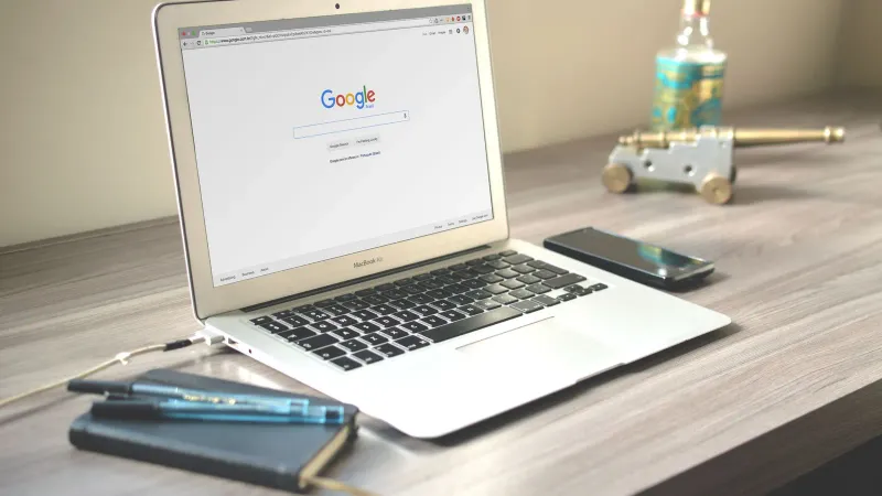 Ein ordentlicher Arbeitsplatz mit einem Laptop, der die Google-Suche anzeigt, einem Smartphone und einem Notizbuch auf einem Holztisch.