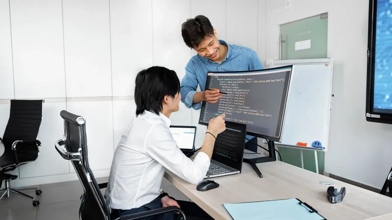 Zwei junge Fachleute arbeiten an einem Schreibtisch mit Laptops und Monitoren in einem modernen Büroumfeld.