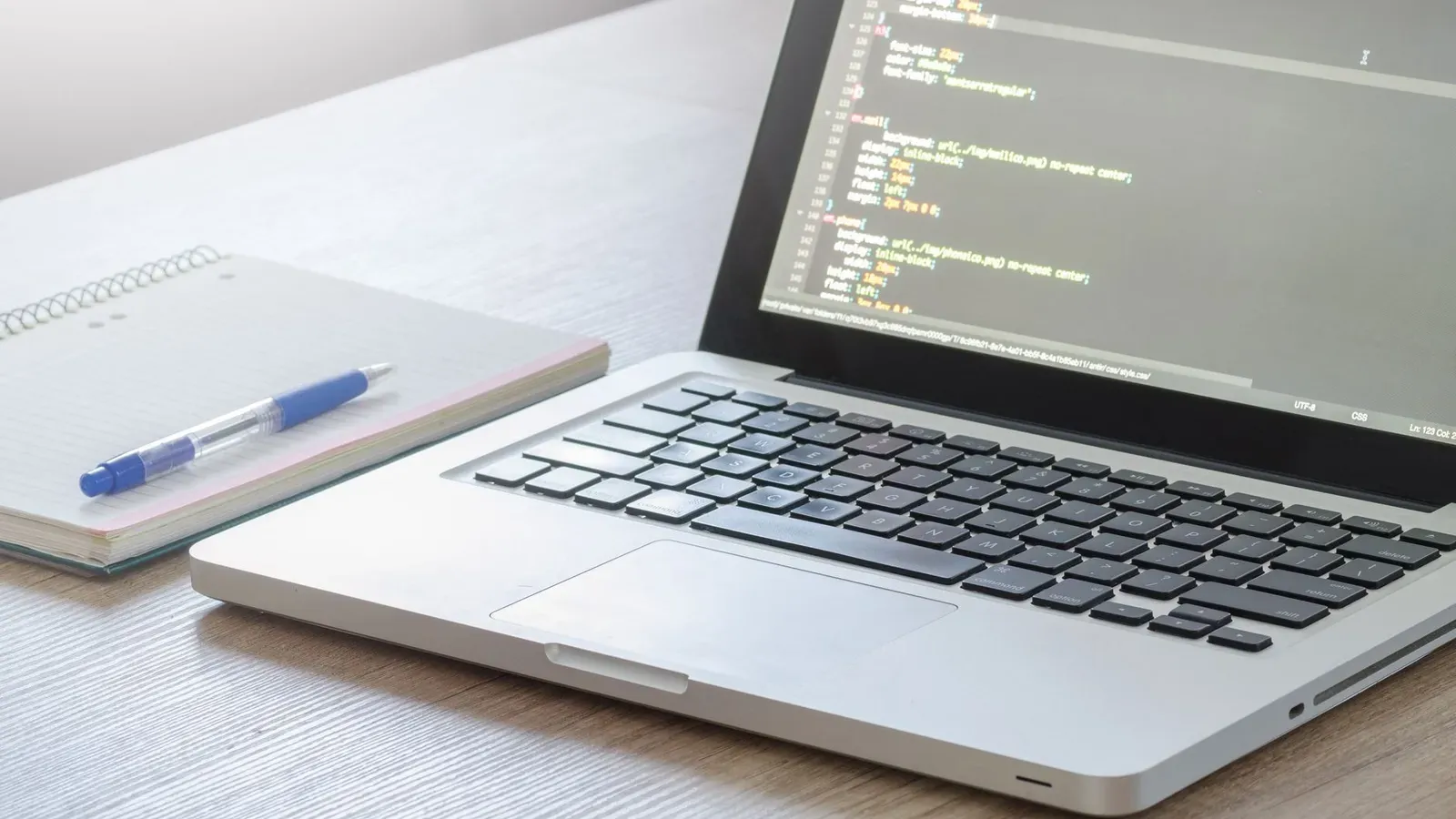 Laptop mit geöffnetem Code-Editor, Notizbuch und Stift auf einem Holztisch, perfekt für Technik- und Arbeitsthemen.