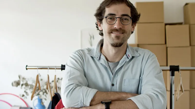 Junger Geschäftsmann mit Brille lächelnd in einem Startup-Umfeld mit Kartons hinter ihm.