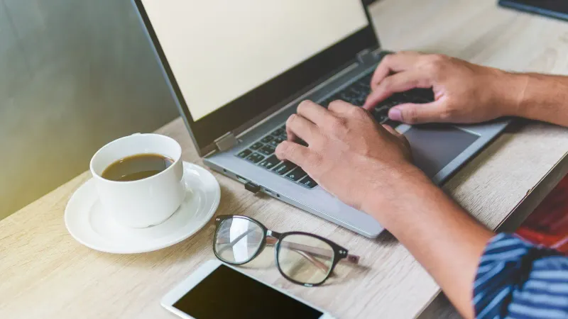 Ein Mann tippt an einem Laptop an einem Schreibtisch mit Kaffee, Telefon und Brille, was ein Homeoffice-Setup veranschaulicht.